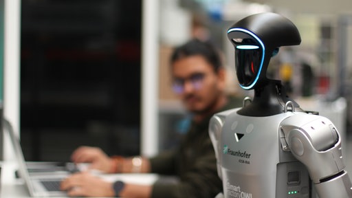 Ein Roboter aus dem Humanoid Robots Experience Lab ist zu sehen, dahinter ein Mitarbeiter des Labors