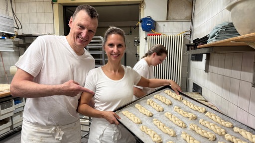 Oliver Güttge und Ann-Kathrin Lüttmann stehen nebeneinander. Sie hält ein Blech mit geflechteten Hefezöpfen in die Kamera.