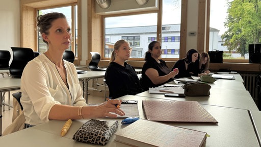 Ann-Kathrin sitzt zusammen mit vier Mitschülerinnen an Tischen in einem Klassenraum und hört dem Lehrer zu.