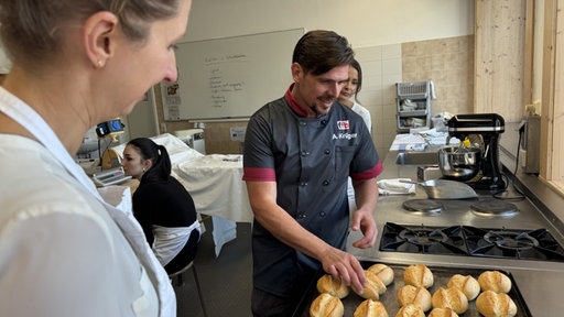 Ann-Kathrin Lüttmann schaut auf ihre gebackenen Weizenbrötchen auf dem Blech. Lehrer Adrian Krüger schaut sie sich an und hält eins in der Hand.