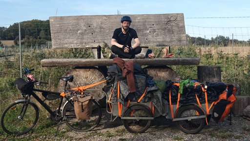Konstantin Wodowos sitz auf einer großen Bank, im Vorderung steht ein Fahrrad mit Anhängern
