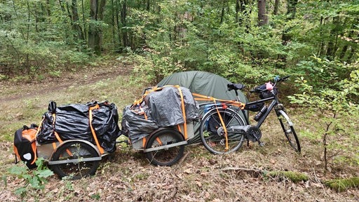 Ein voll beladenes Fahrrad mt Anhängern neben einem Zelt im Wald.