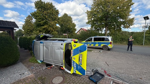 Ein Polizeiwagen liegt umgekippt in einem Vorgarten. Im Hintergrund steht ein Polizist.