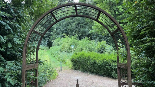 Im Vordergrund ist ein bogenförmiges, verziertes Gartentor aus dünnen Metallstäben zu sehen. Im Hintergrund erstreckt sich ein schmaler Weg, der hinauf in den Wald führt. Auf dem Weg sitzt ein Hase.