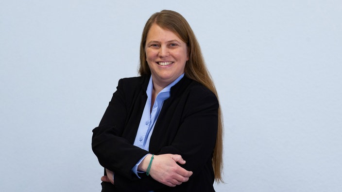 Das Portrait einer Frau mit langen Haaren, sie trägt einen schwarzen Blazer und eine hellbraue Bluse
