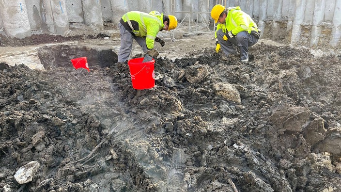 Zwei Archäologen sitzen am Rand der Grube und untersuchen den Inhalt.