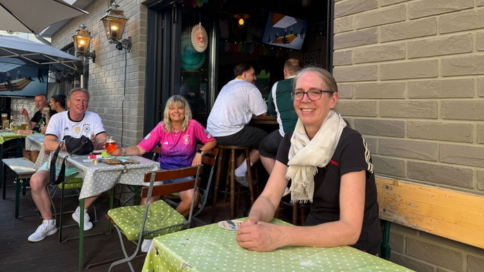 Vor einer Bar sitzen mehrere Gäste an Tischen im Außenbereich und schauen entspannt in die Kamera. Die Atmosphäre wirkt sommerlich und gesellig.
