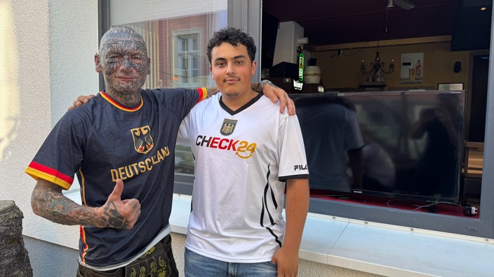 Zwei Fußballfans posieren gemeinsam vor einer Bar für ein Erinnerungsfoto. Beide tragen Deutschland-Trikots und zeigen sich gut gelaunt.