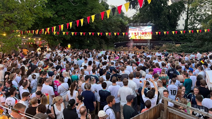Viele Fußballfans verfolgen gemeinsam ein Spiel auf einer Großbildleinwand im Freien. Über der Menschenmenge hängen bunte Wimpelketten in den Farben verschiedener Nationen.