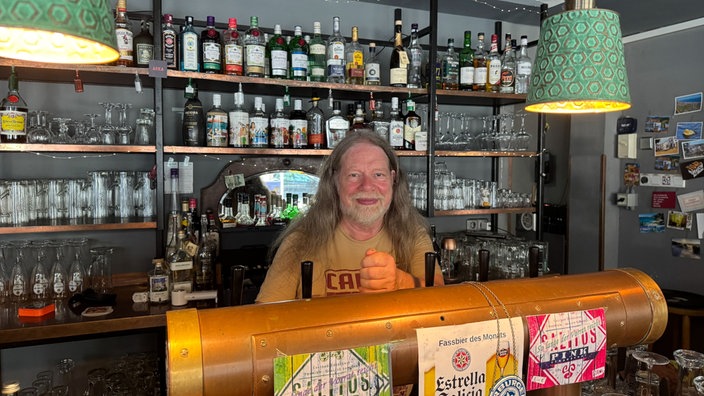 Hinter dem Tresen einer Bar steht ein Mitarbeiter und lächelt in die Kamera. Im Hintergrund sind zahlreiche Flaschen und Gläser zu sehen.