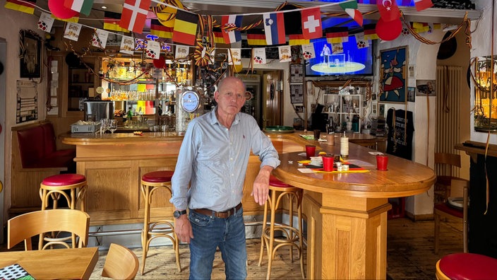 Das Innere einer Fußballkneipe ist mit Trikots, Fahnen und Dekorationen aus verschiedenen Ländern geschmückt. Ein Mann steht im Vordergrund vor der Bar.