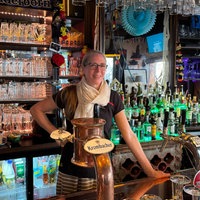 Eine Mitarbeiterin lehnt hinter dem Tresen einer dekorierten Sportsbar und lächelt in die Kamera. Die Bar ist mit vielen Gläsern und Getränken ausgestattet.