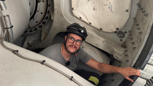 Ein Mann mit Brille und Helm schaut in einem Windrad aus einer Luke und lächtelt in die Kamera