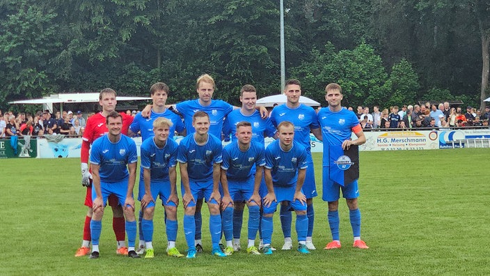 In hellblauen Trikots posieren 11 Fußballspieler im strömenden Regen auf dem Spielfeld.