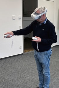 Ein Mann bedient Controller seiner VR-Brille. Im Hintergrund sieht man auf einem Bildschirm, was der Mann mit der VR-Brille sieht.