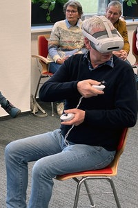 Ein Mann bedient die Controller seiner VR-Brille im Sitzen. Im Hintergrund sitzen andere Menschen und beobachten ihn.