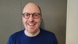 Ein lächelnder Mann mit Glatze und markanter runder Brille, Drei-Tage-Bart und blauem Pulli vor einer grauen Wand.