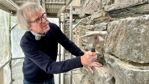 Norbert Sonntag steht neben der Kirchenwand. Er hält einen Hammer in der Hand 