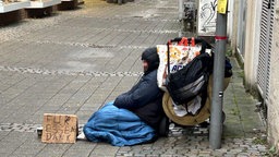 Eine Person sitzt in einem Schlafsack neben einer Schiebewagen und einem Pappschild auf der Straße
