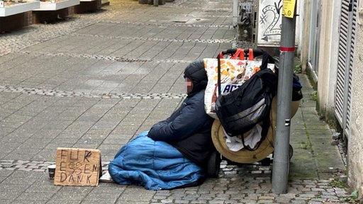 Eine Person sitzt in einem Schlafsack neben einer Schiebewagen und einem Pappschild auf der Straße