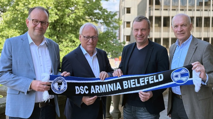 Martin Knabenreich, Pit Clausen, Christoph Wortmann und Rainer Schütte halten einen Arminia-Schal in die Kamera 