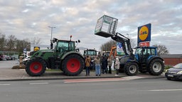 Protest vor einer Lidlfiliale mit Traktoren an denen Schilder befestigt sind. Mehrere Menschen stehen um die Fahrzeugen herum auf dem Lidlparkplatz.