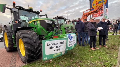 Auf einem Lidlparkplatz: Zwei Traktoren in der Halbfrontale mit Schildern geschmückt auf denen z.B.: "Lebensmittel sind mehr wert" steht. Menschen stehen daneben/dahinter und unterhalten sich.