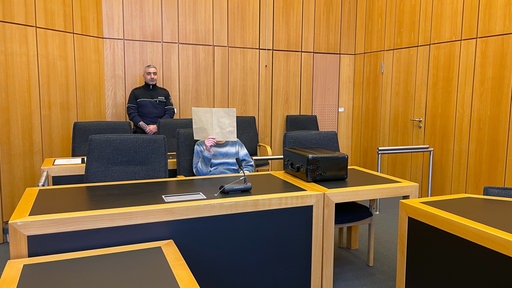 Ein Mann sitzt auf einer Anklagebank in einem Gerichtssaal und hält sich einen Ordner vor das gesicht. Im Hintergrund steht ein Sicherheitsbeamter. 