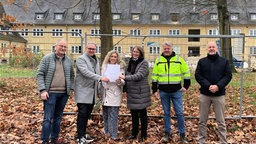 Mehrere Menschen stehen vor einem Bauzaun, im Hintergrund steht das Kasernengebäude