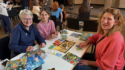 Zwei Frauen und ein Mädchen sitzen an einem Tisch auf dem Spielfelder und Karten mit Pokemons zu sehen sind. Dahinter sind weitere Personen.