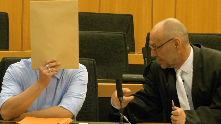 Ein Mann in einem Gerichtssaal mit einem Hefter vorm Gesicht