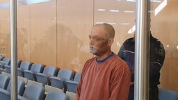 Der Angeklagte steht im Gerichtssaal. Hinter einer Säule steht zur Bewachung ein Wächter.