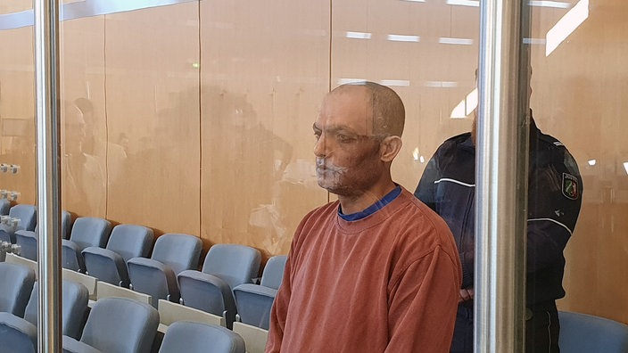 Der Angeklagte steht im Gerichtssaal. Hinter einer Säule steht zur Bewachung ein Wächter.