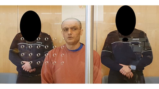 Der Angeklagte steht im Gerichtssaal. Links und rechts hinter ihm stehen zwei Justizbeamte zur Bewachung.