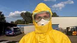 Ein Mann mit Atemschutzmaske, Schutzbrille und gelbem Schutzanzug schaut in die Kamera