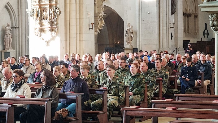 Soldaten in grünen Flecktarn-Uniformen sitzen in einer grauen Steinkirche auf hölzernen Bänken.