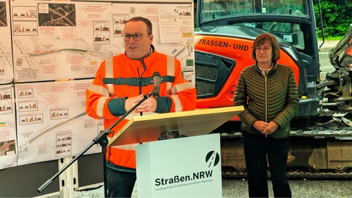 Verkehrsminister Oliver Krischer steht an einem Rednerpult. Hinter ihm sind Pläne zu sehen. Neben ihm steht eine Frau.