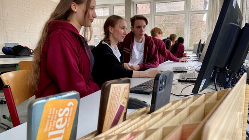 Schülerinnen und Schüler sitzen an Computern und haben Handys vor sich