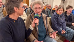 Eine Frau sitzt in einer Kirchenbank und spricht in ein Mikrofon.