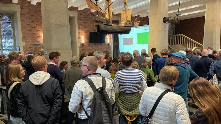 Mehrere Menschen stehen in einem großen Raum. Im Hintergrund ist ein Balkendiagramm auf einer Leinwand zu erkennen. 