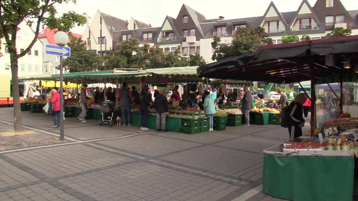 Menschen an mehreren Marktständen auf einem Platz mit umliegenden Gebäuden. 