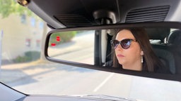 Eine Frau mit Sonnenbrille ist im Rückspiegel eines Autos beim Fahren zu erkennen 