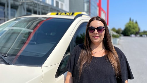 Eine Frau mit Sonnenbrille steht lächelnd vor einem Taxi