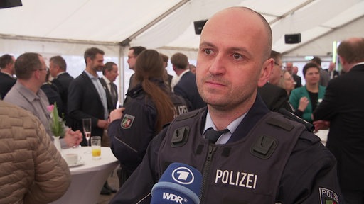 Ein Polizist steht in einem vollen Zelt vor einem Mikro des WDR