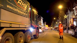 Links im Bild ist ein LKW zu sehen. Rechts von diesem ist eine Person in Wanrweste. Am Straßenrand stehen mehrere Personen. Es ist Nacht. 