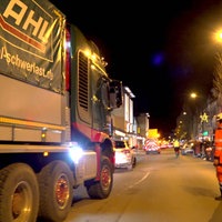 Links im Bild ist ein LKW zu sehen. Rechts von diesem ist eine Person in Wanrweste. Am Straßenrand stehen mehrere Personen. Es ist Nacht. 