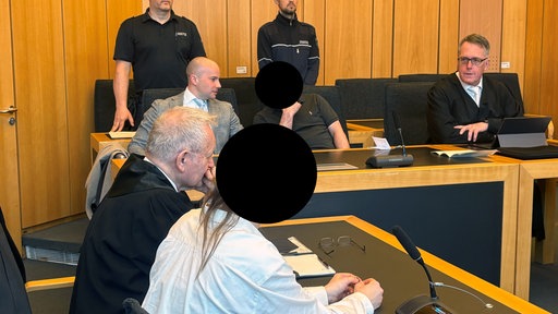 Die zwei Angeklagten sitzen im Gerichtssaal. Daneben sitzen ihre Verteidiger. Hinter ihnen stehen die Gerichtswächter.