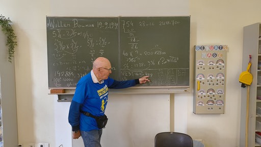 Der Meister im Kopfrechnen, Willem Bouman, steht vor einer Schultafel und zeigt mit seiner linken Hand darauf. Die Tafel ist mit Kreide beschrieben. Auf der Tafel stehen komplexe Brüche, die miteinander multipliziert werden und große Wurzeln.