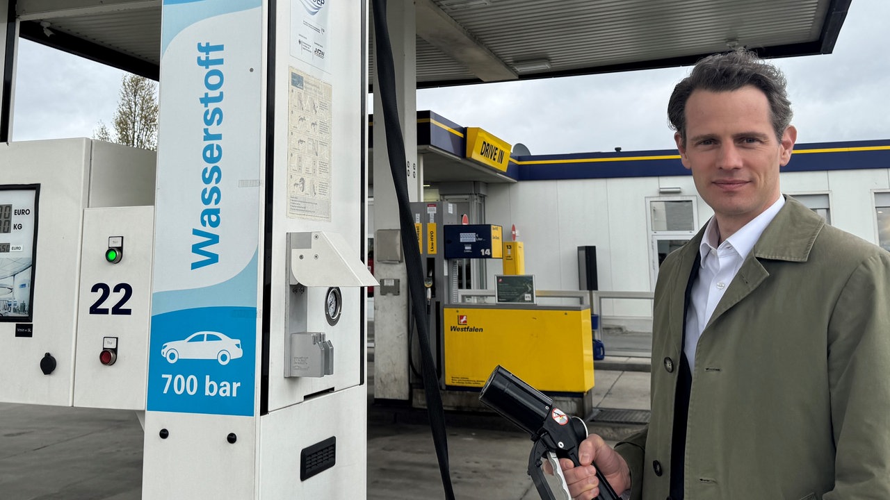 Ein Mann steht neben einer Wasserstoff-Tanksäule und hält einen Zapfhahn in der Hand.