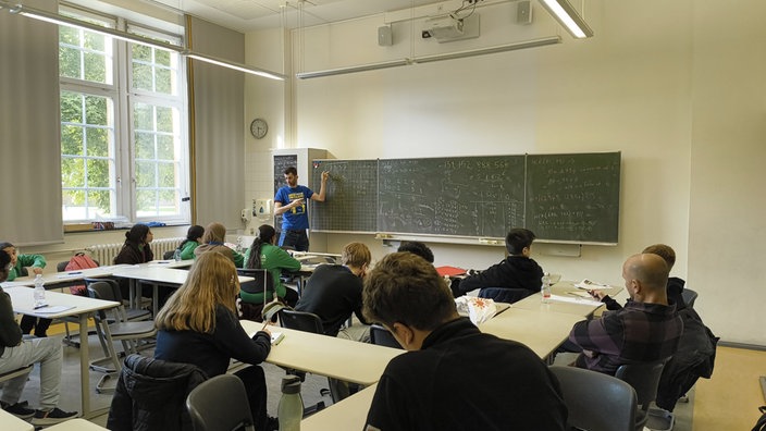 Ein Klassenraum ist zu sehen. Gefüllt ist er mit rund 10 sitzenden Jugendlichen. An der Front des Klassenraums steht Daniel Timms und zeigt auf eine Schultafel. Auf dieser Tafel sind vielstellige Zahlen zu sehen, die miteinander multipliziert werden. 